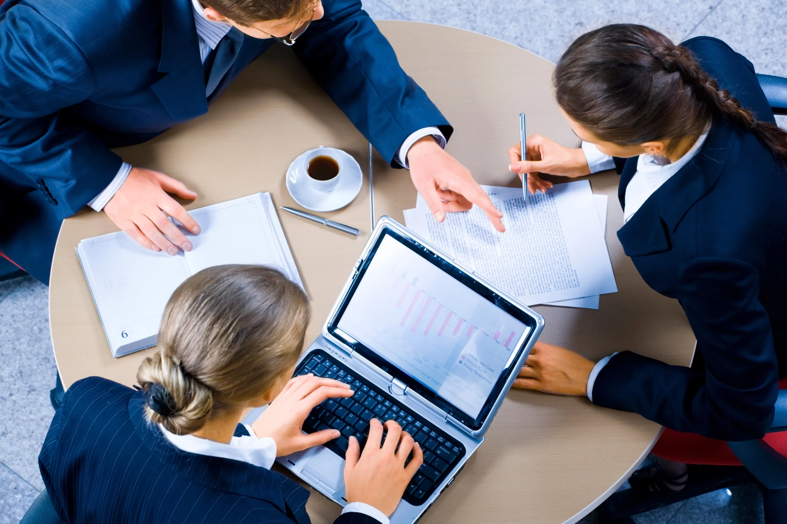 Photo de trois professionnels autour d'une table en train de discuter de données sur un ordinateur portable et des documents imprimés.